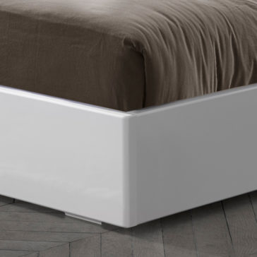Deluxe Miami Style White Lacquered Bed A close-up view of the Deluxe Miami Style White Lacquered Bed with part of the mattress and a dark brown duvet visible. The bed is set on a herringbone-patterned wooden floor. The sleek design features clean lines, creating a minimalist aesthetic.
