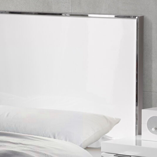 A close-up of the Deluxe Miami Style White Lacquered Bed, featuring a sleek white headboard. The bed is adorned with white linen, and a partially visible white nightstand on the right side of the image holds a small decorative object. The backdrop showcases a light gray wall.