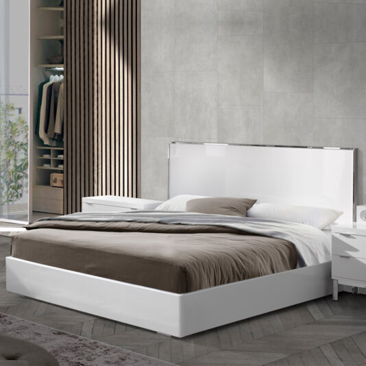 A modern bedroom showcases a Deluxe Miami Style White Lacquered Bed adorned with gray and white bedding. Flanking the bed are a pair of white nightstands. The room highlights gray walls, wooden accents, and an open wardrobe displaying hanging clothes in the backdrop. Sunlight streams through a window, bathing the space in natural light.