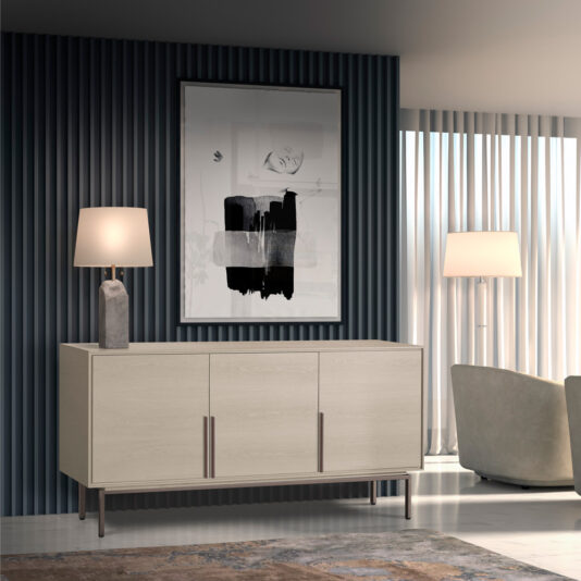A modern living room featuring the Elegant Sideboard With Metal Base, which has three doors and sleek handles, a table lamp with a rectangular ceramic base, and an abstract black-and-white painting above. The floor boasts a patterned rug, and the room is softly illuminated by a tall floor lamp.