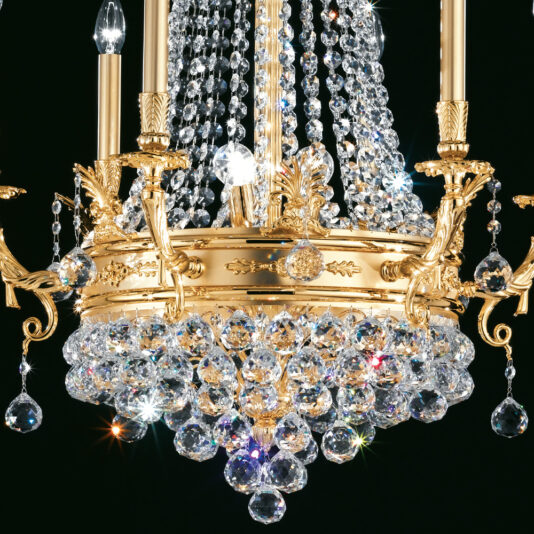 Crystal Drop Empire Chandelier The Crystal Drop Empire Chandelier boasts a gold-finished circular frame, elegantly adorned with multiple crystal strands. Featuring intricate detailing and candle-style light fixtures, the luxurious crystals sparkle brightly, creating a stunning contrast against a dark background.