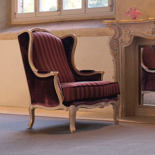 A High Backed Venetian Style Armchair with luxurious burgundy and gold striped upholstery sits next to an ornate fireplace mantel. The chair boasts wooden frames adorned with intricate carvings, and a small decorative fish accent can be seen on the mantel. Light from a window illuminates the entire scene.