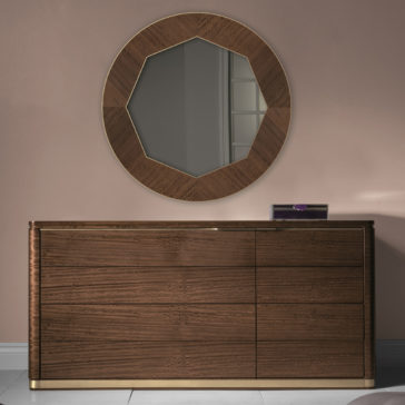 High End Contemporary Chest Of Drawers Product Name: High End Contemporary Chest Of Drawers
Description: The High End Contemporary Chest of Drawers, finished in a luxurious dark brown, stands elegantly against a light pink wall. Mounted above it is an eye-catching large, round mirror featuring a geometric, faceted wooden frame. This exquisite piece of furniture offers three drawers on the right side and is adorned with gold accents at its base.