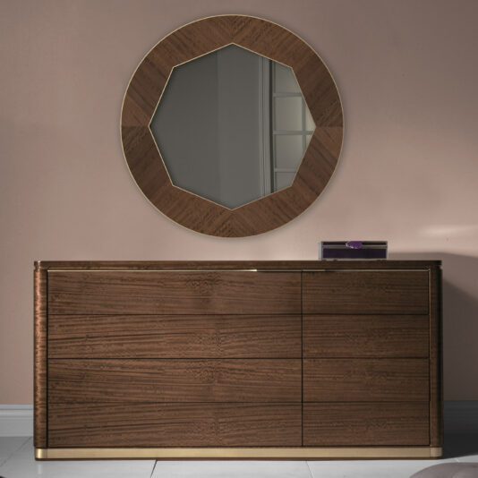 Product Name: High End Contemporary Chest Of Drawers  
Description: The High End Contemporary Chest of Drawers, finished in a luxurious dark brown, stands elegantly against a light pink wall. Mounted above it is an eye-catching large, round mirror featuring a geometric, faceted wooden frame. This exquisite piece of furniture offers three drawers on the right side and is adorned with gold accents at its base.