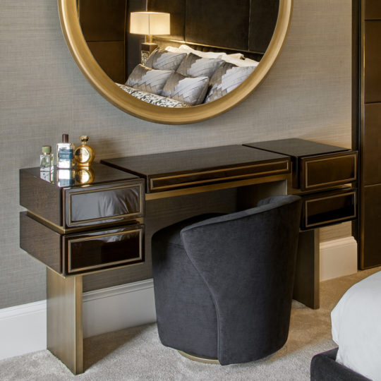 Modern bedroom vanity station featuring the High End Contemporary Dressing Table, complemented by a sleek black chair. The setup includes a sophisticated black and gold desk with multiple drawers, paired with a large round mirror adorned with a gold frame. Placed against a gray wall, the vanity is accessorized with toiletries and has a lamp that is reflected in the mirror.