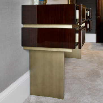 High End Contemporary Dressing Table A close-up of the High End Contemporary Dressing Table against a gray wall. The dresser features sleek, dark wood drawers adorned with white handles, and is supported by gold-colored, rectangular legs. The floor is carpeted in light beige, enhancing the contemporary and minimalist style of the space.