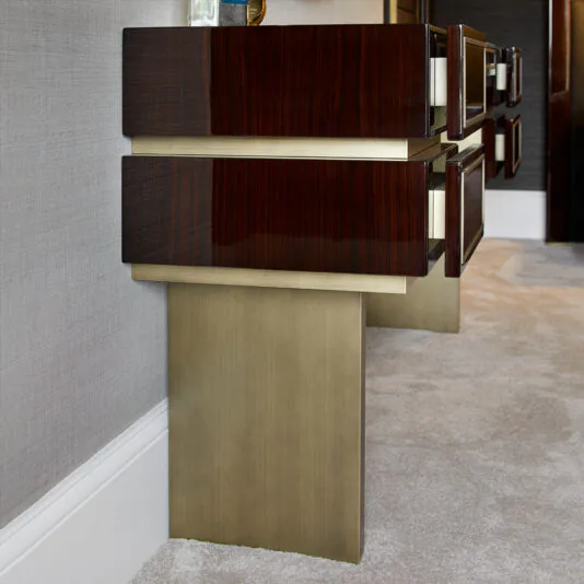 High End Contemporary Dressing Table A close-up of the High End Contemporary Dressing Table against a gray wall. The dresser features sleek, dark wood drawers adorned with white handles, and is supported by gold-colored, rectangular legs. The floor is carpeted in light beige, enhancing the contemporary and minimalist style of the space.