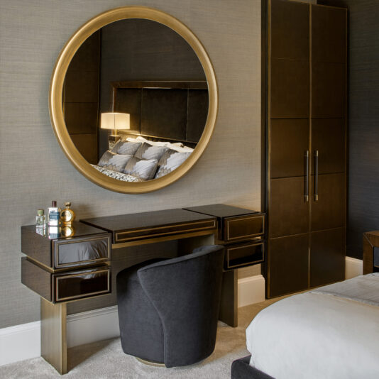 High End Contemporary Dressing Table A modern bedroom features the High End Contemporary Dressing Table, showcasing a sleek vanity paired with a large, round, gold-framed mirror. The dark wood vanity includes gold accents and three drawers. Positioned in front is a matching plush chair. Nearby, a tall wardrobe and bed are visible, complemented by an elegant table lamp beside the bed.