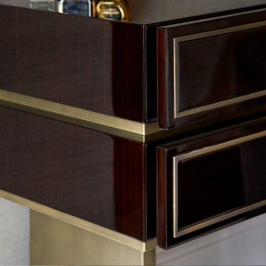 High End Contemporary Dressing Table A close-up of the High End Contemporary Dressing Table reveals its modern, glossy wooden surface with a dark finish and gold accents. The image zeroes in on the upper corner, highlighting detailed craftsmanship and sleek design elements such as a partially visible drawer and some small items placed on the table's surface.