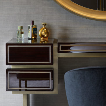 High End Contemporary Dressing Table A High End Contemporary Dressing Table with dark wood and gold accents, featuring two drawers. Positioned on top are four perfume bottles of various shapes and colors. Partially visible above the table is a large round mirror with a gold frame, and a dark gray upholstered chair is in view.