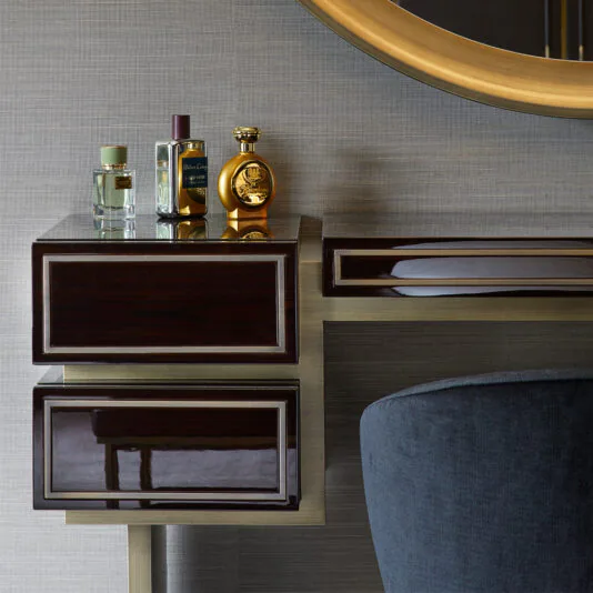 High End Contemporary Dressing Table A High End Contemporary Dressing Table with dark wood and gold accents, featuring two drawers. Positioned on top are four perfume bottles of various shapes and colors. Partially visible above the table is a large round mirror with a gold frame, and a dark gray upholstered chair is in view.