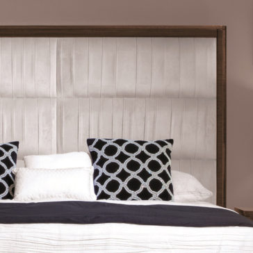 Contemporary Pleat Upholstered Bed A section of an upscale bedroom features a **Contemporary Pleat Upholstered Bed** with a large padded beige headboard, neatly arranged white and dark blue bedding, and decorative pillows with a linked circle pattern in blue. The background has a light brown wall.