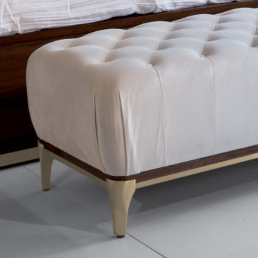 High End Contemporary Ottoman Bench A close-up photograph of the High End Contemporary Ottoman Bench showcasing its white, tufted cushion and wooden base and legs, placed on pristine white tiled flooring. In the background, part of a wooden bed frame and a crisp white bedspread on the mattress are visible.