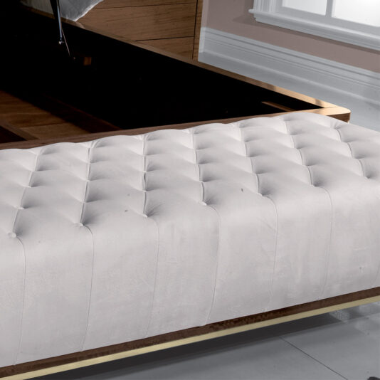A close-up of a high-end contemporary bedroom showcases a tufted white headboard with button details. The bedframe, crafted from dark wood and boasting a shiny finish, is set against a wall painted in a light pastel color. In the background, a large window provides ample natural light, while at the foot of the bed sits the High End Contemporary Ottoman Bench.