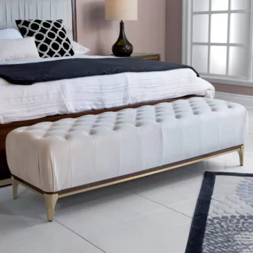 High End Contemporary Ottoman Bench A chic bedroom showcases the elegant Luxury Button Upholstered Stool with gold legs positioned at the foot of a bed. The bed is dressed in sleek white and black bedding, complemented by a nearby bedside table that holds a stylish black lamp. Natural light pours through a large window, accentuating the modern decor and tiled flooring throughout the space.