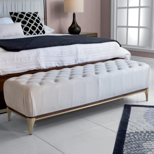 A chic bedroom showcases the elegant Luxury Button Upholstered Stool with gold legs positioned at the foot of a bed. The bed is dressed in sleek white and black bedding, complemented by a nearby bedside table that holds a stylish black lamp. Natural light pours through a large window, accentuating the modern decor and tiled flooring throughout the space.