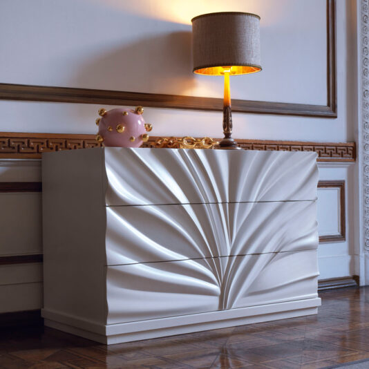The High End Modern Chest Of Drawers, featuring a unique wavy design on the drawer fronts, stands against an ornate wall with wood paneling. On top of the dresser are a lit lamp with a brown shade and a decorative pink and gold orb. The floor showcases a polished wooden surface.