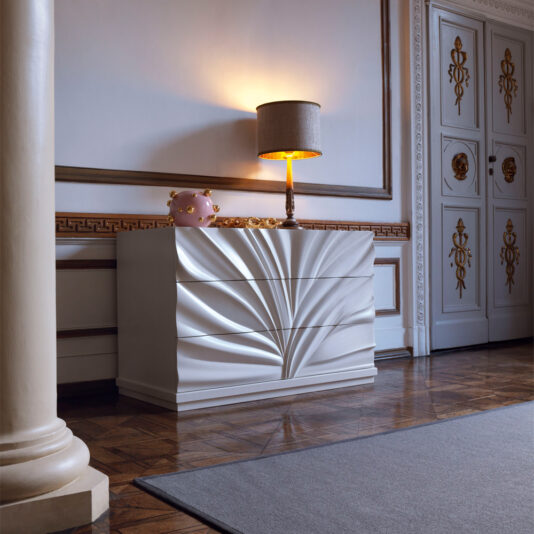 High End Modern Chest Of Drawers A luxurious room featuring the High End Modern Chest Of Drawers, adorned with an ornate white design that showcases artistic, fan-like patterns on the drawers. Atop the chest is a lit lamp with a beige shade and a pink piggy bank. The floor boasts a patterned wooden design, while elegant white walls with gold accents surround the space.