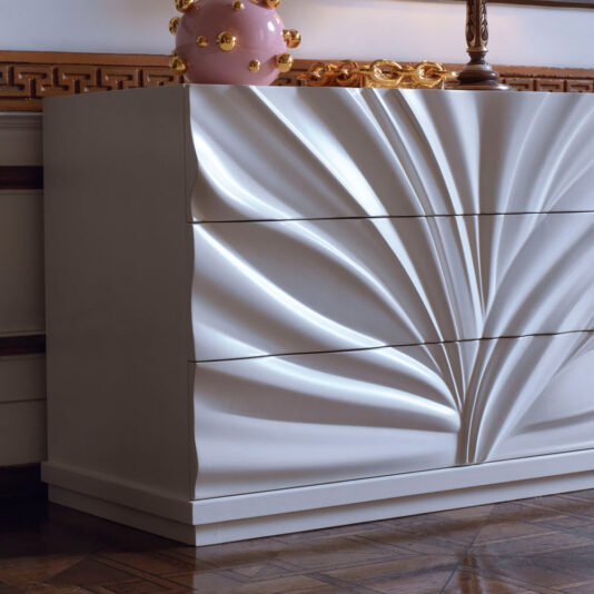 High End Modern Chest Of Drawers The High End Modern Chest Of Drawers features a sophisticated, flowing wave-like design on its drawers, combined with a minimalist aesthetic. It stands elegantly on a wooden floor, and a decorative piece with pink and gold accents graces the top.