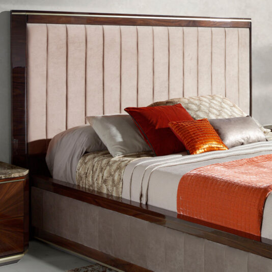 The High End Modern Upholstered Bed showcases a sophisticated upholstered headboard with vertical panels, framed gracefully by polished wood. The bed is adorned with a variety of pillows in shades of grey, white, and vibrant red, accompanied by a neatly folded burnt orange quilt. A side table can be partially seen beside the bed.