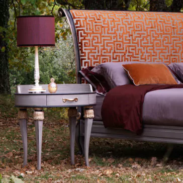 High End Oval Bedside Cabinet With Drawer A luxurious bed with an orange and gold patterned headboard and burgundy linens is set outdoors among trees. Next to the bed on the grass stands a High End Oval Bedside Cabinet With Drawer, adorned with intricately designed legs and holding a vintage-style lamp along with decorative items.