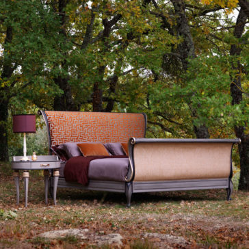 High End Oval Bedside Cabinet With Drawer A stylish bed with an ornate headboard and footboard featuring geometric patterns is situated outdoors among green trees. The High End Oval Bedside Cabinet With Drawer, paired with a purple lamp and a small circular table adorned with decorative ornaments, adds elegance to the scene. Plush pillows and bedding cover the bed.