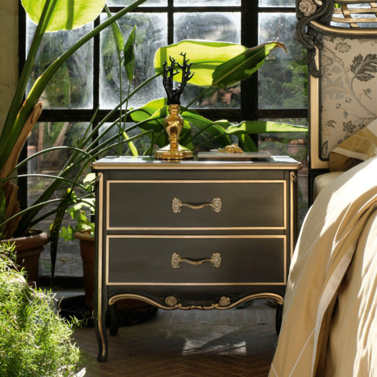 The Large Classic Ornate Bedside Cabinet with two drawers and ornate handles is positioned next to a large window. The cabinet supports a gold decorative candle holder, while sunlight streams through the window, illuminating surrounding indoor plants and a cozy, fabric-covered bed.