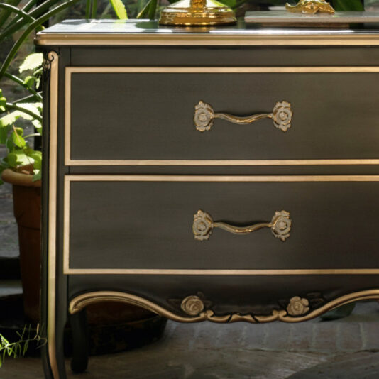 A close-up of the Large Classic Ornate Bedside Cabinet, showcasing its elegant black and gold finish with two spacious drawers. The top drawer is adorned with handles shaped like decorative leaves, while the bottom drawer features intricate floral accents. An ornate golden object is partially visible on the cabinet's surface.