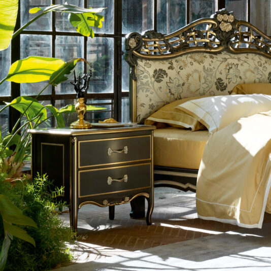 A decorative bedroom featuring a vintage floral-patterned headboard bed, yellow bedding, and the Large Classic Ornate Bedside Cabinet with two spacious drawers. The room is filled with abundant natural light streaming through large windows and is adorned with lush green plants, creating a serene ambiance.