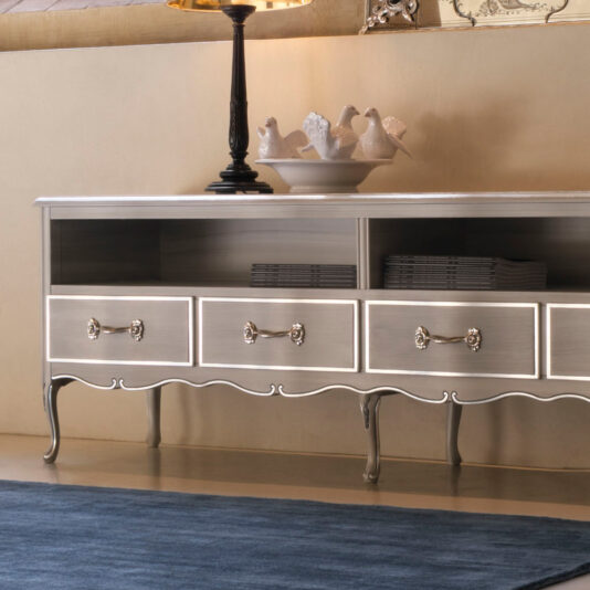 Large Classic Style TV Unit With Drawers A Large Classic Style TV Unit With Drawers stands against a light-colored wall. It features a silver, vintage-style finish with curved legs and ornate drawer handles. Atop the unit, there is a decorative bowl with ceramic birds. Below the tabletop are open shelves containing neatly stacked books and a partially visible lamp.