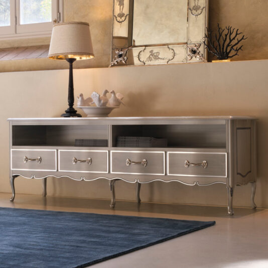 A stylish, metallic Large Classic Style TV Unit With Drawers stands against a beige wall. Resembling a sideboard with ornate detailing, it features two open shelves and six drawers with decorative handles. A lamp and several decorative items are placed on top, while a large, elegant mirror is mounted on the wall behind it.