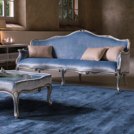 A stylish living room features the Large Classic Venetian Style Sofa in blue velvet with silver trim and two beige cushions. In front of the sofa is an elegant, matching coffee table. The room is decorated in muted tones with a large window providing natural light.