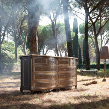 A Large Double Louis Style Chest Of Drawers sits in a sunlit forest clearing. Tall trees and scattered sunlight create a serene atmosphere, enhanced by a lamp and bench in the background.