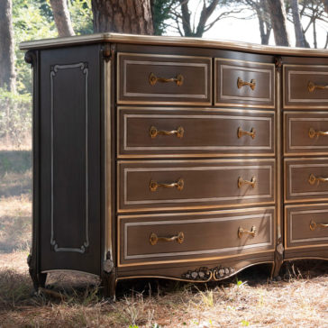 A Large Double Louis Style Chest Of Drawers with ornate brass handles and intricate detailing stands outdoors. Sunlight filters through the surrounding trees, casting a warm glow on the vintage-style wooden chest. Featuring multiple drawers with decorative trim and elegantly curved legs, it exudes classic charm.