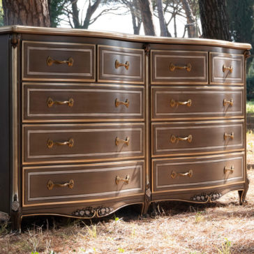 The Large Double Louis Style Chest Of Drawers is a beautifully crafted piece with an elegant design, featuring nine drawers adorned with ornate golden handles. This double dresser boasts a rich wood finish and is set outdoors amidst tall trees, with sunlight filtering through the branches.