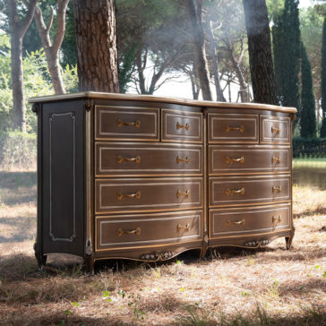The Large Double Louis Style Chest Of Drawers, featuring ornate brass handles and decorative carvings, stands outdoors in a forested area. Sunlight filters through the trees, casting a soft light on the dresser while the ground is covered with dry pine needles.