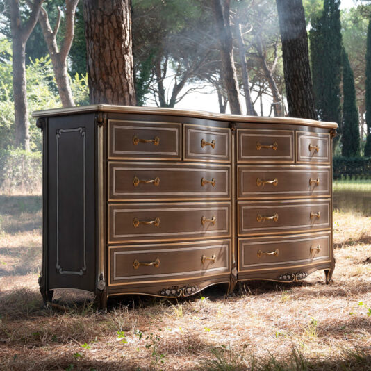 The Large Double Louis Style Chest Of Drawers, featuring ornate brass handles and decorative carvings, stands outdoors in a forested area. Sunlight filters through the trees, casting a soft light on the dresser while the ground is covered with dry pine needles.
