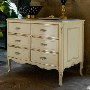 A vintage-style cream-colored Louis Style Chest Of Drawers featuring six drawers, ornate handles, and decorative carvings stands against a stone wall. The chest is adorned with a black bowl with white accents and a gold tray holding miscellaneous items, while a large green plant is partially visible in the background.