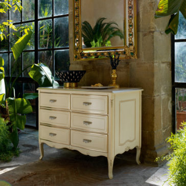 A vintage Louis Style Chest Of Drawers in cream stands against a large window in a bright, indoor greenery-filled space. It features six drawers and is topped with a gold-bordered mirror, a decorative bowl, and a candelabra. The setting is lush with various green plants and natural light.