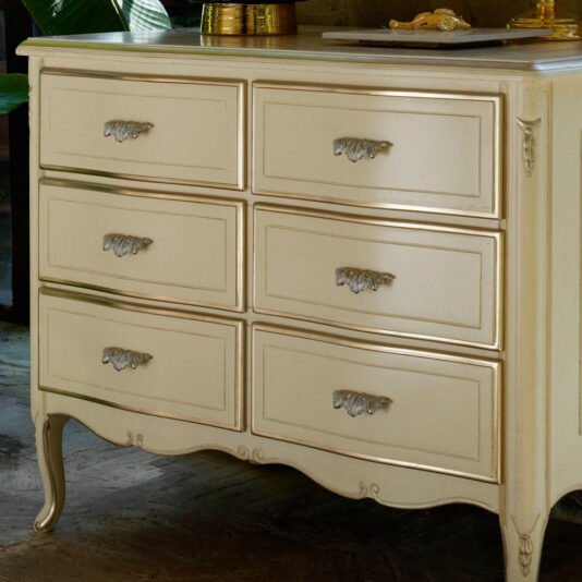 Louis Style Chest Of Drawers The Louis Style Chest Of Drawers is a vintage-inspired piece, cream-colored and featuring six drawers with ornate silver handles. Its curved legs and decorative edging along the sides exude classic elegance. Placed on a wooden floor, the top surface is adorned with a few items, including a small gold vase.