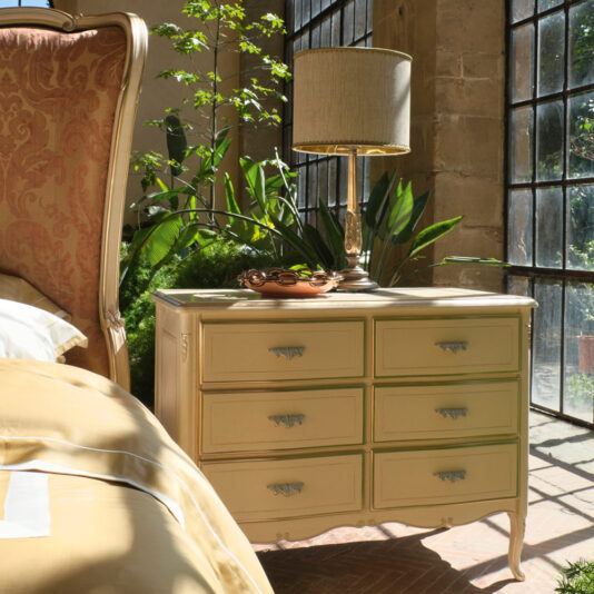 Louis Style Chest Of Drawers A sunlit bedroom features a beige upholstered bed with a nightstand alongside it. The nightstand, similar to the Louis Style Chest Of Drawers, has six drawers and intricate handles. It is adorned with a beige table lamp and a small tray, surrounded by lush green plants. Large windows bring in natural light.