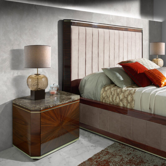 A modern bedroom featuring a dark wooden headboard with vertical padding, a neatly made bed with beige and red bedding, two Luxury Dark Rosewood Bedside Tables topped with elegant lamps, a soft, textured gray wall in the background, and a patterned rug on the floor.