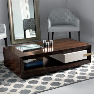 A modern living room with a wooden coffee table featuring shelves and a white drawer. On the table are a book, a magazine, and three candles. Behind the table are a large mirror and a gray upholstered chair. The floor has a patterned rug with gray tones.