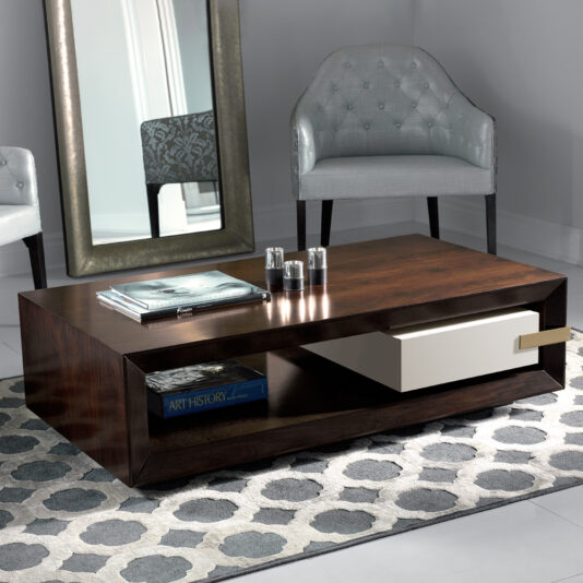 A modern living room with a wooden coffee table featuring shelves and a white drawer. On the table are a book, a magazine, and three candles. Behind the table are a large mirror and a gray upholstered chair. The floor has a patterned rug with gray tones.