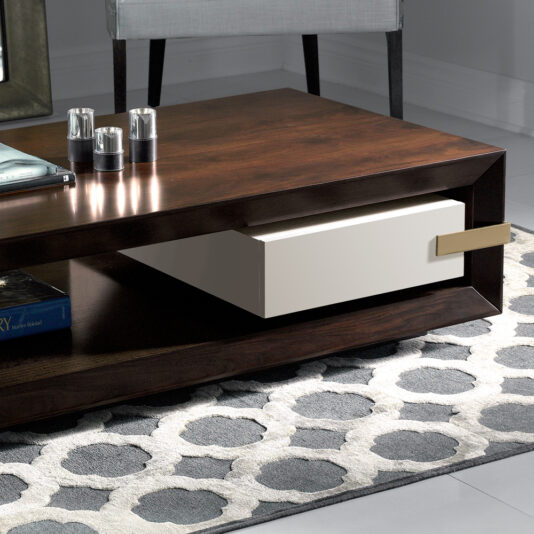 A modern wooden coffee table with a white drawer and metallic handle, positioned in a contemporary living space. The table sits on a patterned gray and white rug. There are candle holders and a book placed on the table.