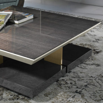 Modern Veneer Square Coffee Table A Modern Veneer Square Coffee Table with a dark gray marbled top and gold accents is positioned in a stylish living room. Books are neatly placed on the table, which sits on a plush, patterned gray rug. The table boasts a sleek, geometric base design.