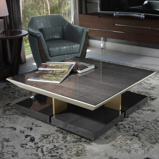 A modern living room with a comfortable blue armchair and a glass-topped coffee table featuring gold accents. The Modern Veneer Square Coffee Table, adorned with books, adds to the chic ambiance. In the background, a wooden sideboard and large windows allow natural light to flow in.