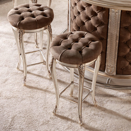 Two **Luxury Italian Bar Stools** with tufted brown velvet seats and ornate light-colored legs placed on a plush beige carpet evoke the charm of opulent dressing table stools. The bar behind them features a matching tufted design, creating a cohesive and sumptuous setting.