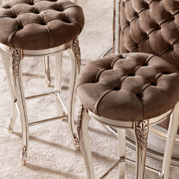 Luxury Italian Bar Stool A close-up of two Luxury Italian Bar Stools with plush, tufted brown cushions and intricately carved silver legs, placed on a light beige carpet. Part of a matching padded and tufted surface, reminiscent of a luxurious Italian dressing table stool, is visible in the background.