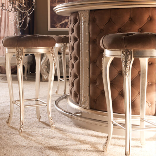 Luxury Italian Bar Stool A close-up view of an elegant bar area featuring tall Luxury Italian Bar Stools with tufted brown leather and ornate metallic legs, complemented by a matching bar with tufted leather siding. The setting exudes a warm, luxurious atmosphere with soft lighting and lavish decor, reminiscent of an opulent Italian dressing table stool.
